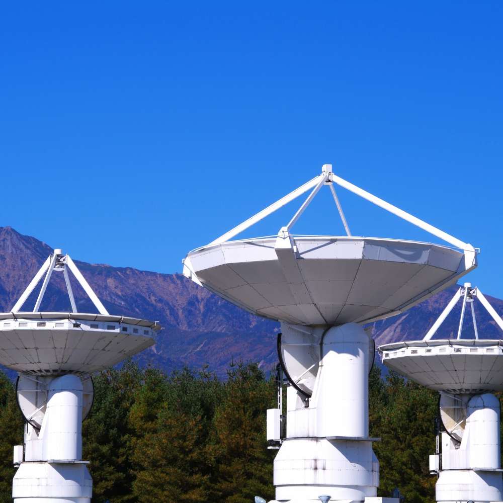 Large white radio telescope dishes at an observatory with forest and mountain background under a clear blue sky.