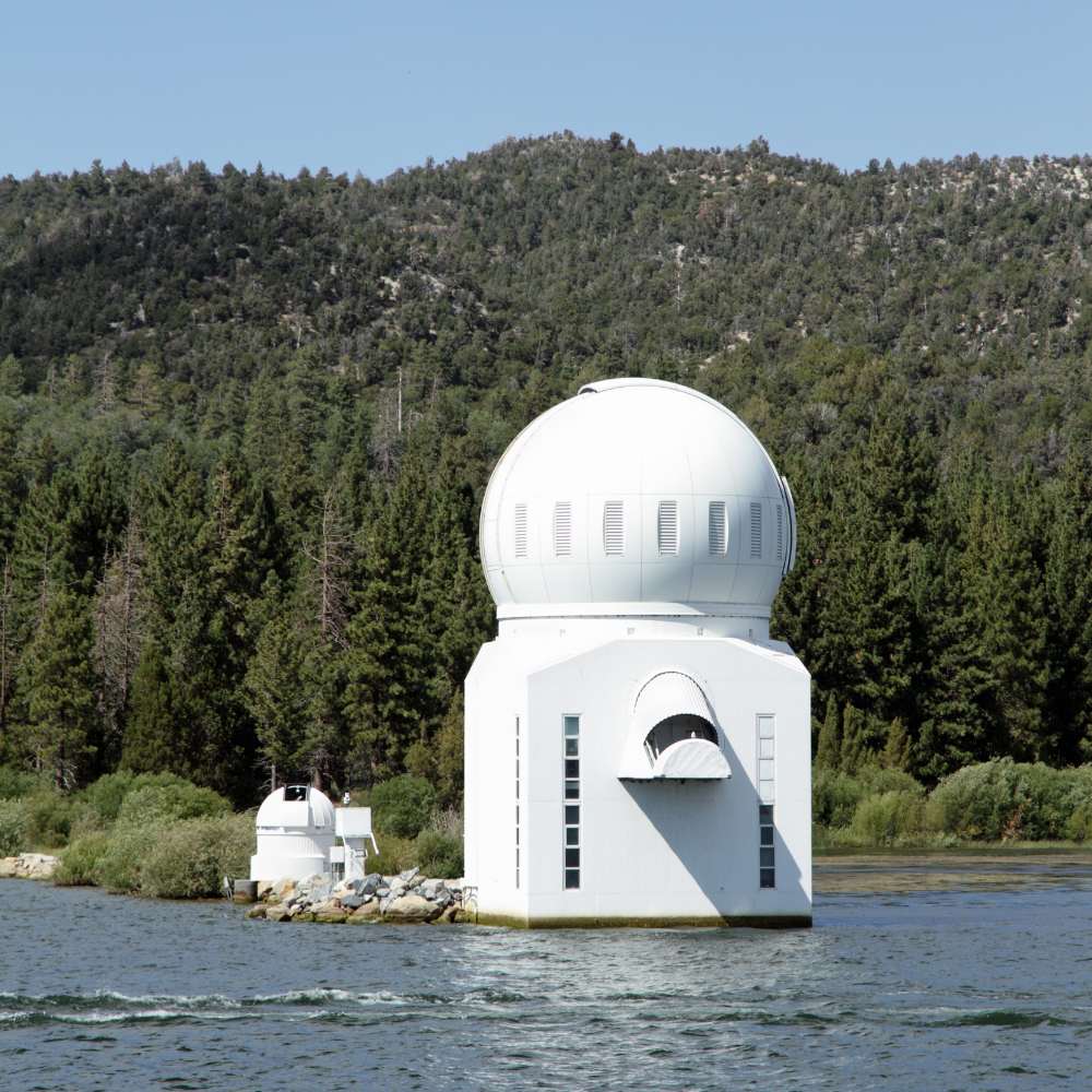 White solar observatory dome built on a lake, surrounded by dense pine forest and mountains under a clear sky.